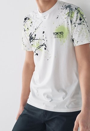 Maglietta bianca con design a schizzi multicolori in nero e verde neon, con testo stampato "TOKYO" e coordinate.