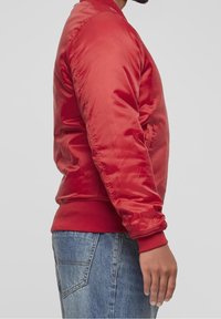 Giacca bomber rossa realizzata in tessuto liscio, con polsini e orlo a coste, tasche laterali e design aderente. Indossata sopra jeans blu.
