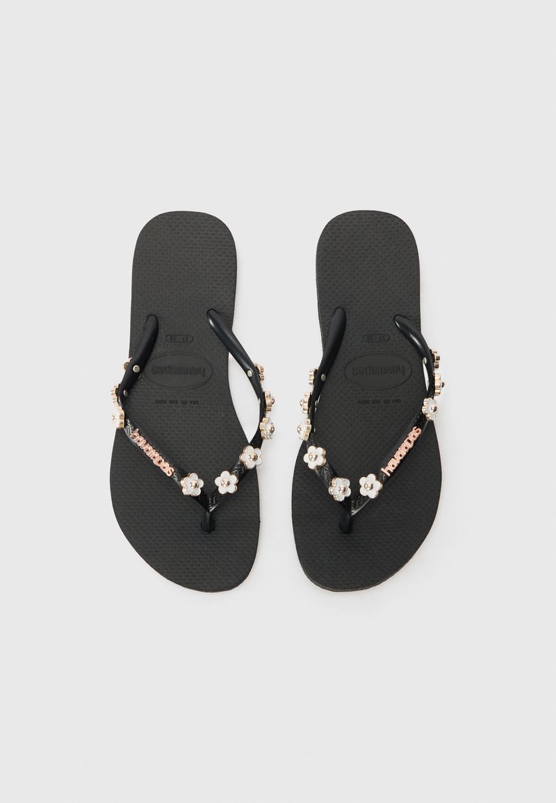 Μαύρες ελαστικές παντόφλες με υφή στο πέλμα, διακοσμημένες με λουλουδάτο χάντρα και λεπτομέρεια λογότυπου στη λουρίδα. Ελαφρύ και casual σχέδιο.