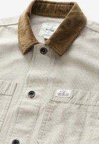 Chemise à rayures beige et blanche avec col en velours côtelé marron, poches avant et une étiquette cousue sur la poche poitrine gauche.
