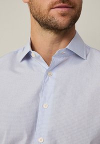 Camicia a scacchi azzurro chiaro con colletto button-down, caratterizzata da una sottile texture e quattro bottoni bianchi sul davanti.