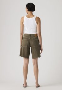 Γυναίκα που στέκεται πλάτη, φοράει λευκό αμάνικο τοπ, ελιάς πράσινα σορτς με φθαρμένο τελείωμα και μαύρα σανδάλια slide σε απλό φόντο.