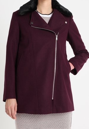 Femme portant un manteau en laine violet foncé avec un col en fausse fourrure noire, une fermeture éclair argentée inclinée et les mains dans les poches latérales.