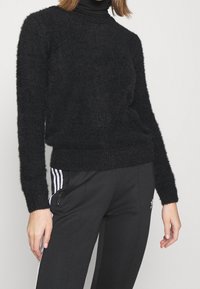 Kobieta w puszystym czarnym sweterze z długim rękawem na czarnym golfie, w połączeniu z czarnymi spodniami dresowymi Adidas z białymi paskami.