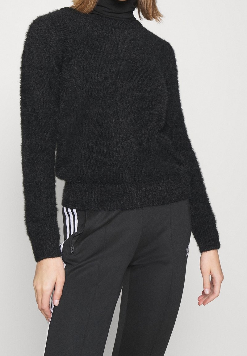 Kobieta w puszystym czarnym sweterze z długim rękawem na czarnym golfie, w połączeniu z czarnymi spodniami dresowymi Adidas z białymi paskami.