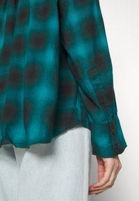 Personne portant une chemise à carreaux bleu sarcelle et noire à manches longues avec poignets boutonnés, associée à un pantalon gris clair sur un fond blanc uni.