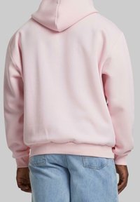 Person som bär en ljusrosa hoodie och ljusblå jeans, sedd bakifrån mot en enfärgad grå bakgrund.