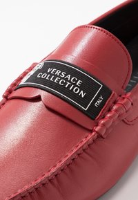 Rote Leder-Mokkasins mit einem schwarzen Stofflogo-Patch, das "VERSACE COLLECTION" liest, und geflochtenen Details entlang der Kanten. Glatte Textur.