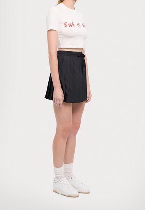 Kvinde iført hvid crop-top med rød tekst, sort plisseret nederdel, hvide sokker og hvide sneakers, stående foran en ensfarvet baggrund.