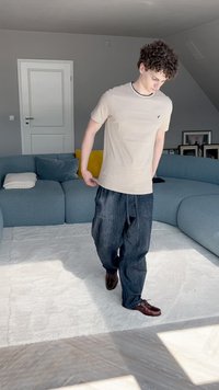 Jasny beżowy t-shirt z krótkim rękawem i czarnymi wykończeniami, w połączeniu z luźnymi, ciemnoniebieskimi spodniami cargo z denimu oraz brązowymi butami na sznurówki, w jasnym pomieszczeniu.