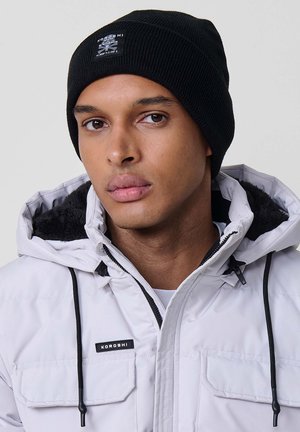 Gorra negra de punto con textura acanalada y un pequeño parche de logo. Lleva puesta una chaqueta blanca con aislamiento y capucha forrada de piel sintética.