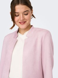Jonge vrouw met een lichtroze blazer over een wit shirt, zacht glimlachend en naar beneden kijkend tegen een lichte achtergrond.