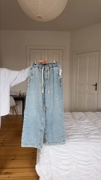 Svijetloplave široke hlače od trapera s elastičnim pojasom i vezicom, koje drži osoba u svijetloj spavaćoj sobi s drvenim podom i bijelim prekrivačem na krevetu.