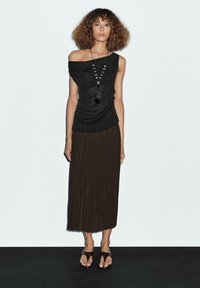 Donna con i capelli ricci che indossa un top nero a spalle scoperte, una lunga gonna plissettata nera, una collana di perline nera e sandali neri, in piedi contro un muro bianco.
