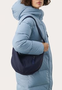 Navyblaue Stoff-Schultertasche mit runder Form und Reißverschluss, getragen über einem hellblauen Puffermantel mit großer Kapuze.