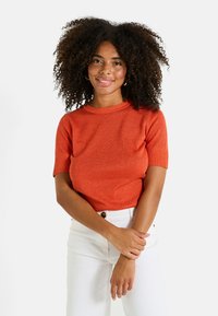 Suéter de punto de canalé de manga corta en naranja brillante con cuello redondo. Combinado con pantalones blancos, que cuentan con un detalle de botón.