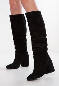 Svarta mocka knähöga stövlar med slouchig skaft och klackar, bärs på nakna ben mot vit bakgrund.