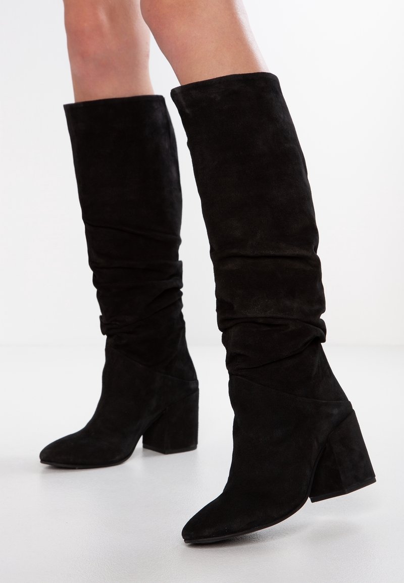 Svarta mocka knähöga stövlar med slouchig skaft och klackar, bärs på nakna ben mot vit bakgrund.