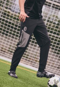 Zwarte sportbroek met grijze accenten, ritszak aan de zijkant en logo op de dij, gecombineerd met zwarte voetbalschoenen, op een veld.