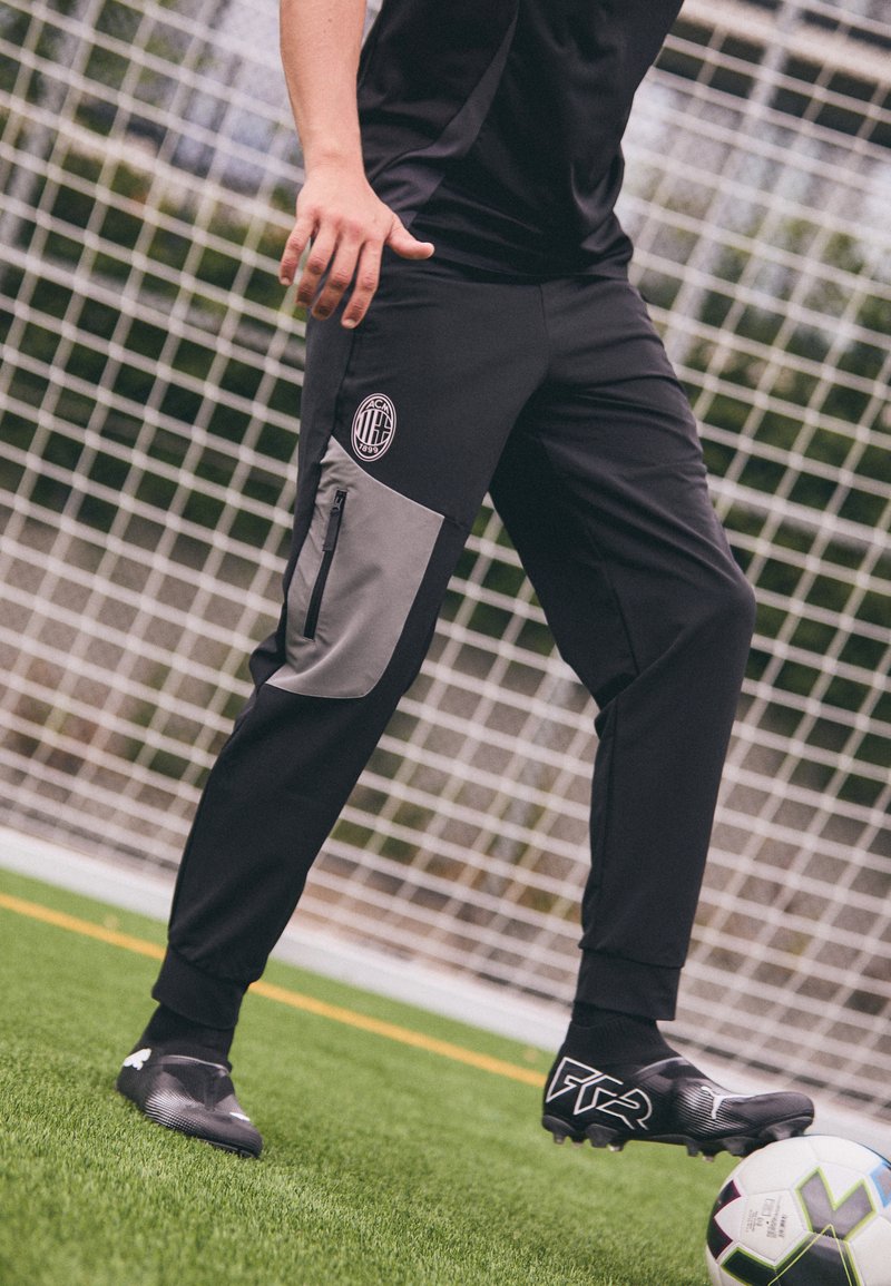 Zwarte sportbroek met grijze accenten, ritszak aan de zijkant en logo op de dij, gecombineerd met zwarte voetbalschoenen, op een veld.