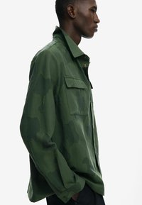 Green camo shirt with a structured collar, front pocket, and raw-edge details. Made of lightweight fabric with a textured finish.