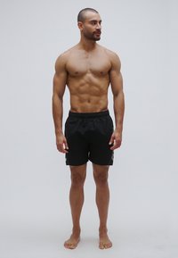 Pantalones cortos de baño negros con cintura elástica, con detalles grises en los lados. Modelo está de pie descalzo sobre un fondo liso.