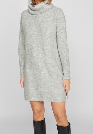 Frau trägt ein hellgraues, langärmliges Strickkleid mit lockerem Rollkragen, kombiniert mit schwarzen kniehohen Stiefeln.