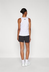 Hvitt, ermløst treningstopp med logo på ryggen, kombinert med svarte shorts og hvite joggesko; glatt stoff og sporty design.