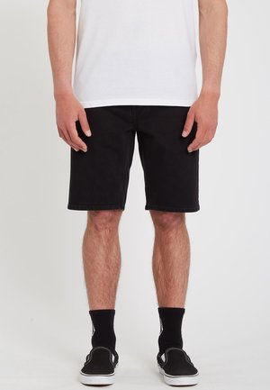 Mann trägt schwarze Shorts, schwarze Socken, schwarze Slipper und ein weißes T-Shirt und steht vor einem einfarbigen Hintergrund.