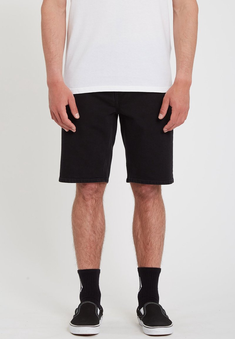 Homme portant un short noir, des chaussettes noires, des chaussures plates noires et un T-shirt blanc, debout devant un fond uni.
