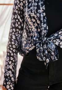 Transparente dunkelblaue Bluse mit leicht metallic glänzendem Blumenmuster, an der Taille gebunden. Darunter sind schwarze hochtaillierte Jeans sichtbar. Weicher, strukturierter Stoff.