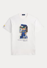 Λευκό T-shirt με γραφικό του Polo Bear ντυμένο με μπλε αθλητικά ρούχα, κρατώντας τσάντες, που στέκεται δίπλα σε τρένο με κείμενο "Polo Bear by Ralph Lauren."