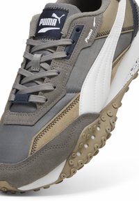 Grauer und beiger Puma-Sneaker mit weißem Streifen, strukturiertem Gummisohle mit Noppen und grauen Schnürsenkeln, schräg ausgerichtet, um Vorder- und Seitendetails zu zeigen.