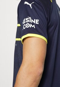 Tummansininen urheilupaita lyhyillä hihoilla, jossa on keltaisia yksityiskohtia, teksturoitu muotoilu ja valkoinen brändäys hihassa.