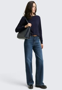 Frau trägt einen marinefarbenen Pullover, weite blaue Jeans, schwarze Schuhe, hält eine große schwarze Schultertasche und steht vor einem schlichten hellen Hintergrund.