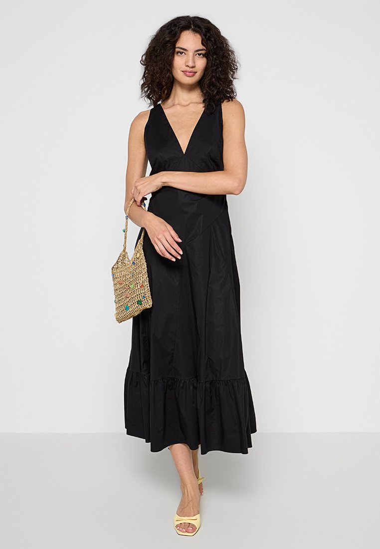 Kobieta z kręconymi włosami, ubrana w bezrękawową czarną sukienkę midi, trzymająca plecioną torbę ze słomy i żółte sandały na obcasie z odkrytymi palcami.