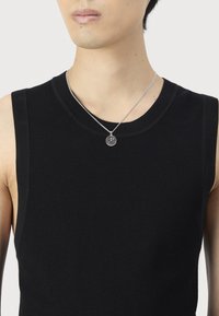 Persoană îmbrăcată cu un top negru fără mâneci din tricot și un colier dintr-un lanț argintiu cu un pandantiv rotund, pe fundal alb.