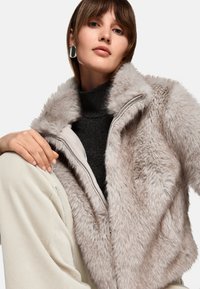 Faux-Fur-Jacke in Hellgrau mit Reißverschluss, übergroßem Kragen und strukturiertem Finish. Kombiniert mit einem schwarzen Rollkragenpullover und hellbeigen Hosen.