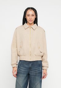 Casaco cropped bege com fecho de correr, gola grande e dois bolsos frontais. Apresenta detalhes dourados e punhos canelados nas mangas.