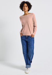 Helles rosa Sweatshirt mit weitem Ausschnitt und gerippten Bündchen, kombiniert mit blauen Jeans und weißen Sneakern. Lässiger, lockerer Schnitt.