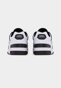 Ζευγάρι λευκά και μαύρα χαμηλά αθλητικά παπούτσια, ορατά από πίσω, με μαύρες σόλες και επενδεδυμένους πάτους, σε απλό φόντο.