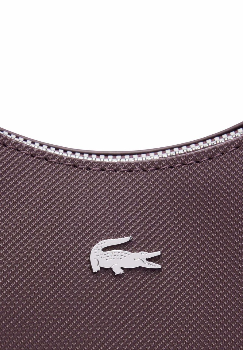 Nahaufnahme eines braunen strukturierten Stoffes mit einem silbernen Krokodil-Logo über einer Reißverschlussnaht.