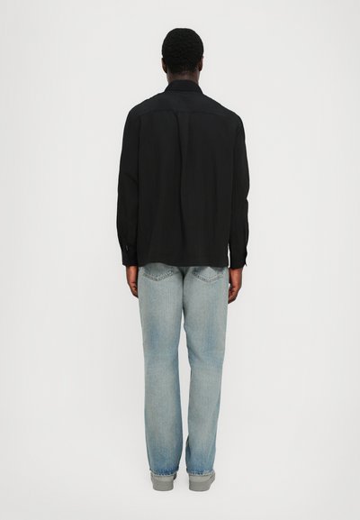 Mustalane varrukatega särk struktureeritud silueti ja nööpidega mansettidega, millel on tagumised voldid, koos helesiniste sirgete pükste ja hallide jalatsitega.