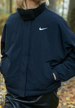 Osoba ubrana w granatową kurtkę Nike z czarnym kołnierzem z materiału typu sherpa, z rękami w kieszeniach, stojąca na zewnątrz wśród jesiennych liści.