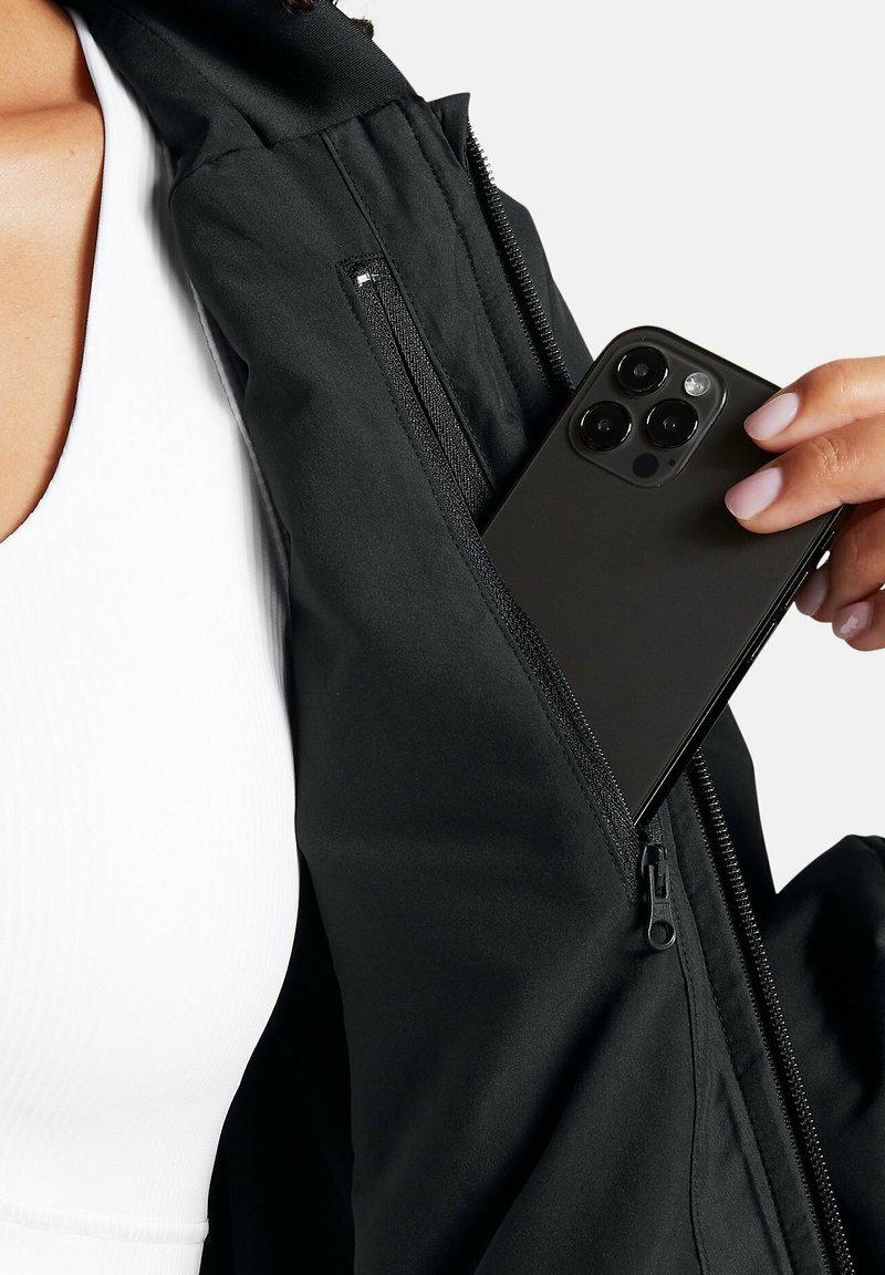 Czarna kurtka z widoczną kieszenią na zamek błyskawiczny, w której znajduje się ciemny smartfon. Tkanina wydaje się gładka, o matowym wykończeniu. Pod spodem biały top.