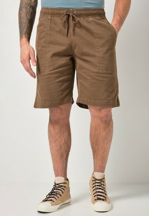 Mann trägt braune Shorts mit Kordelzug und beige High-Top-Sneaker, steht mit den Händen in den Taschen vor schlichtem Hintergrund.