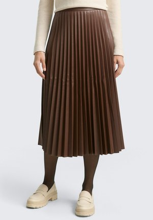 Frau trägt einen braunen plissierten Midirock, schwarze Strumpfhosen, cremefarbene Loafers und ein beiges langärmeliges Oberteil, stehend vor einem einfarbigen Hintergrund.