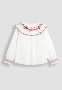 Weiße Langarmbluse mit einem Rüschenausschnitt, versehen mit bunten, floralen Stickereien. Die Manschetten haben smockiertes pinkes Detail. Strukturierter Stoff.
