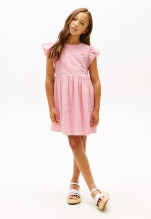 Ragazza giovane in piedi con gambe incrociate, indossa un vestito a righe rosa e bianco e sandali piattaforma bianchi su uno sfondo semplice.