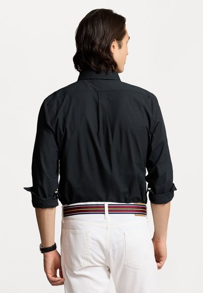Svart skjorta med knappar och uppvikt ärm, bakfold, som bärs med vita byxor och ett flerfärgat randigt bälte. Minimalistisk design.